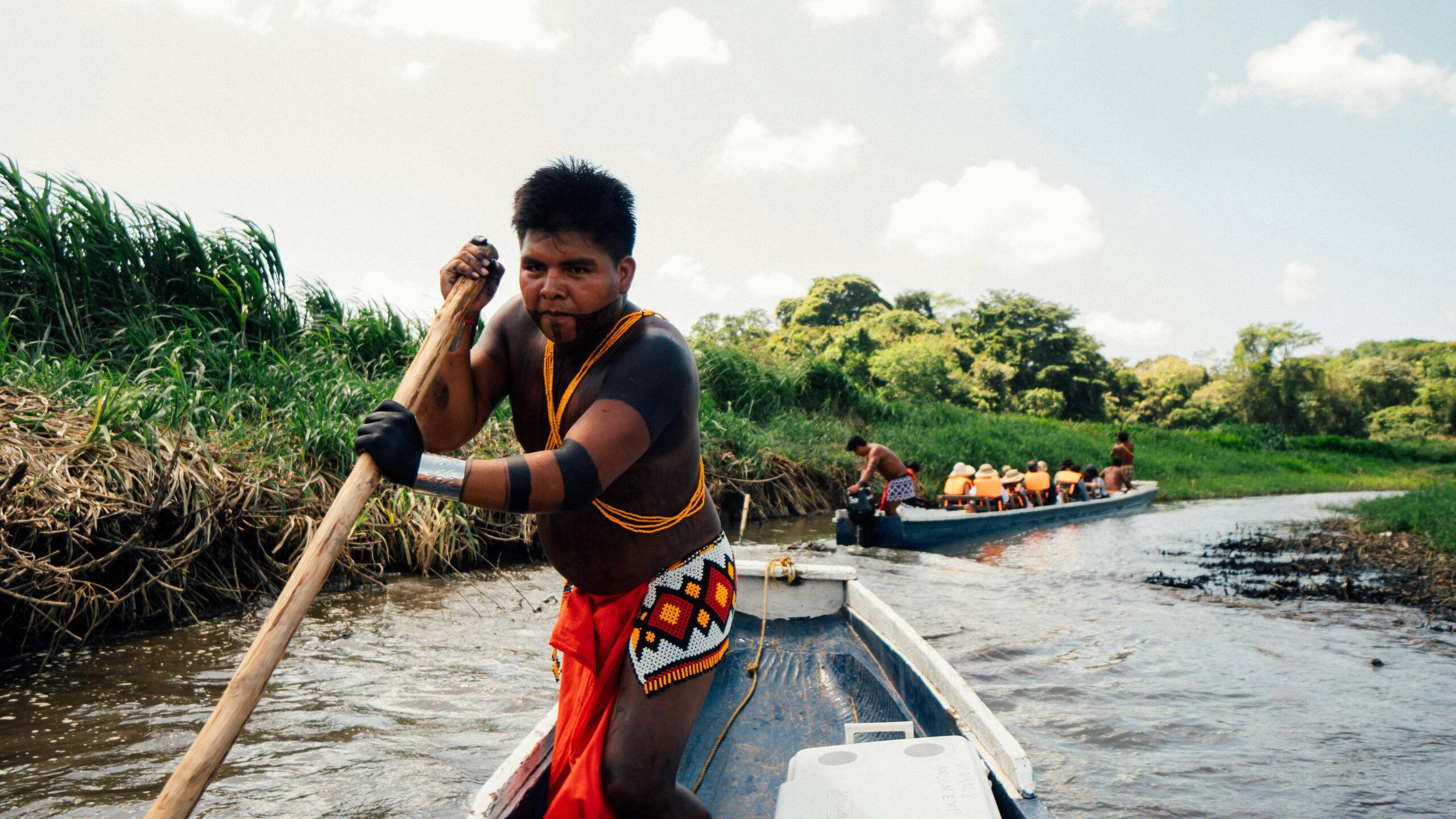 Indigenous people canoeing in traditional wear on a river through lush greenery during daytime.